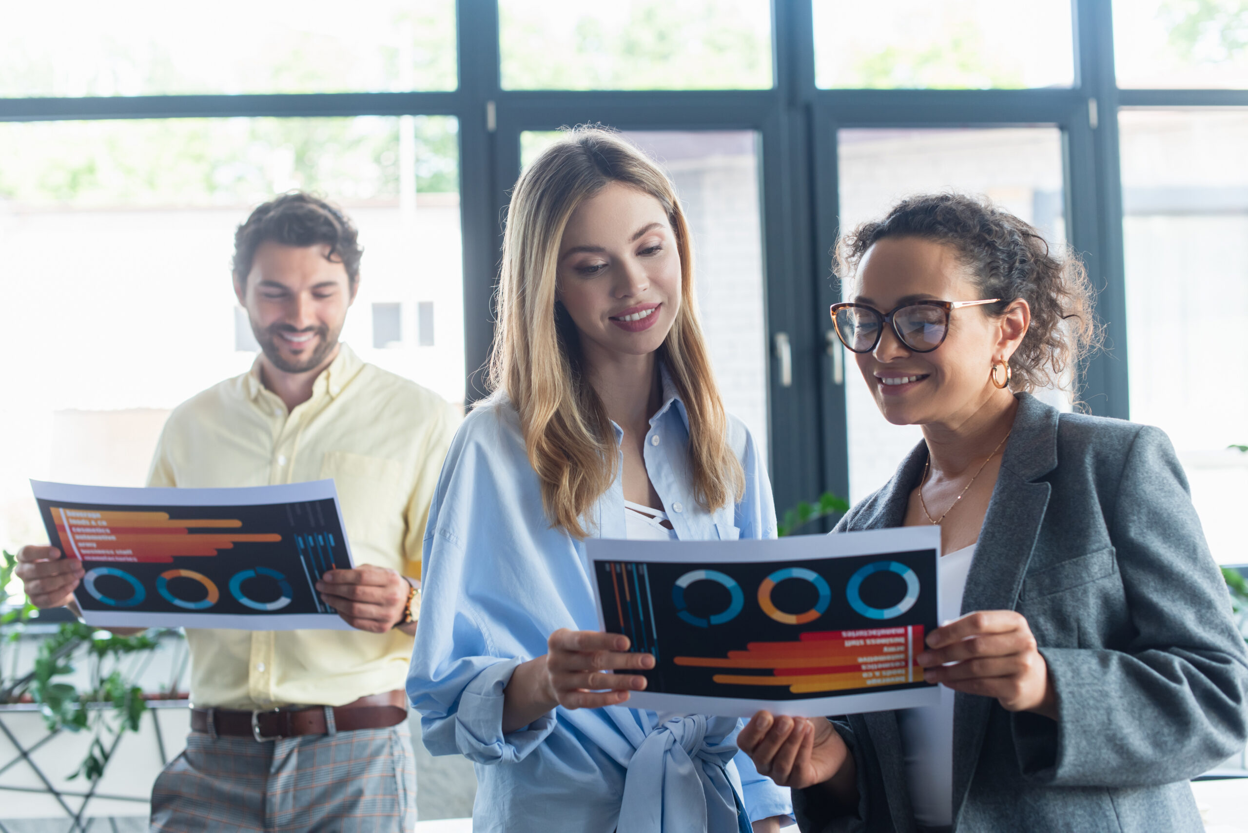 Smiling SaaS Sales multiethnic businesswomen holding SaaS document near blurred colleague in office